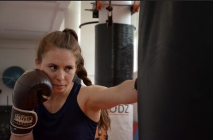 Zdjęcie przedstawia dziewczynę w rękawicach bokserskich która uderza w worek treningowy