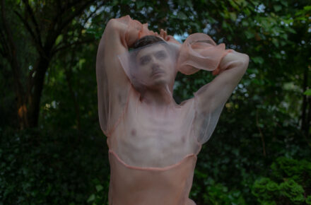 Zdjęcie przedstawia osobę AMAB z rękoma podniesionymi do góry i ubraną w prześwitujący, sitakowy kostium